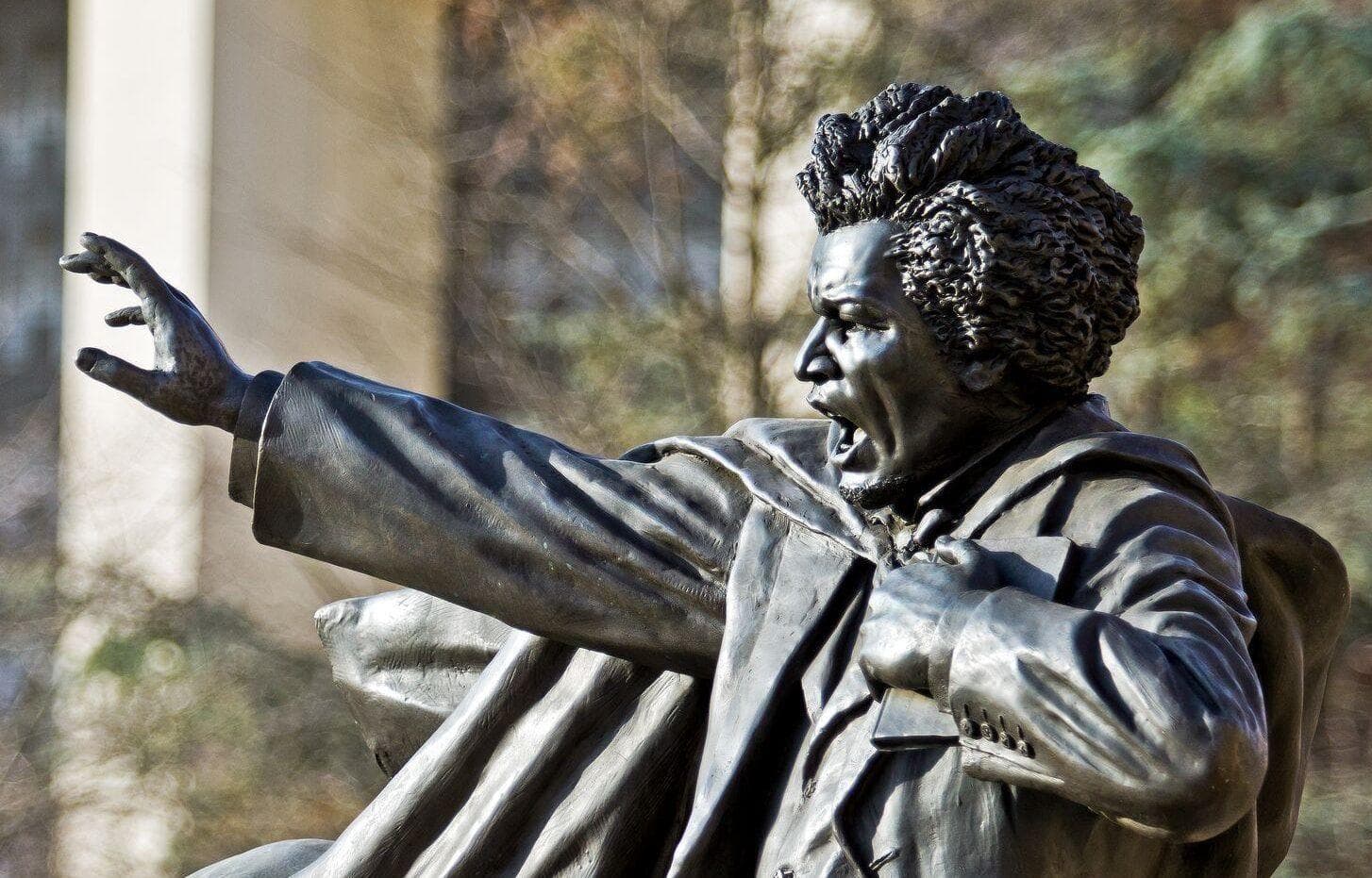 Weathered bronze statue of abolitionist, social reformer and statesman, Frederick Douglass. His right arm raised while delivering a powerful speech. His left arm holds tight a copy of his autobiography.